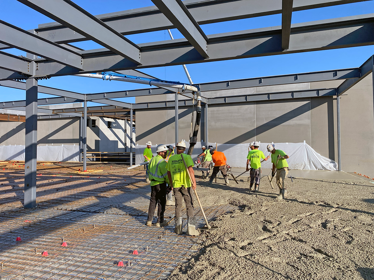 Construction site with a steel frame, workers in hard hats and bright vests spreading and smoothing wet concrete over a rebar grid under a clear blue sky.